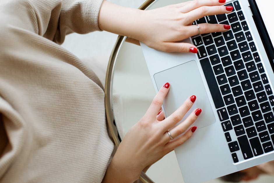 A woman with red nails typing on a laptop, view from above, indoors setting.