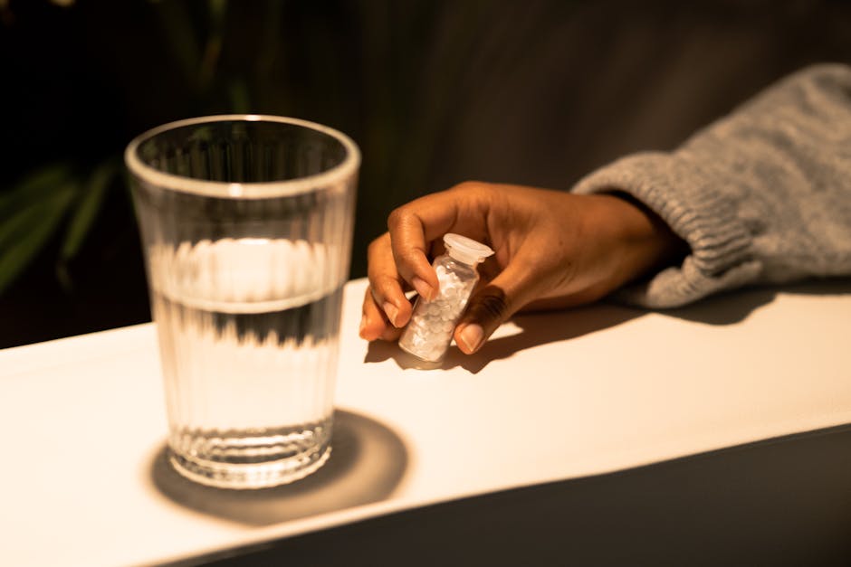 Close-up of hand holding a pill container beside a water glass, symbolizing health and medication.