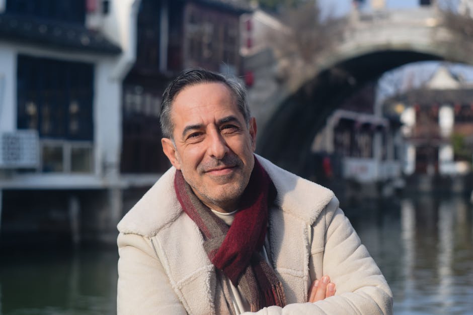 A middle-aged man smiles confidently near a canal bridge, wearing a winter coat and scarf.