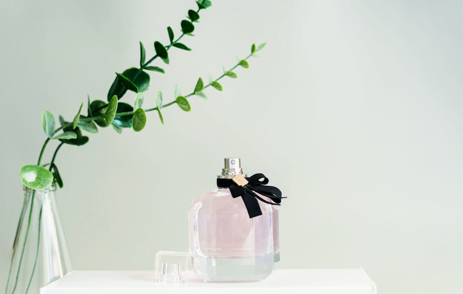 A close-up of a perfume bottle with a green eucalyptus branch in a vase on a white background.