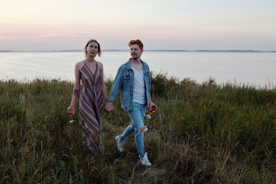A couple holding wine glasses walks hand in hand by a serene lakeside at sunset, capturing a romantic mood.