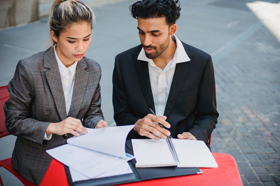 Two professionals in business suits reviewing documents while sitting outdoors at a red table.