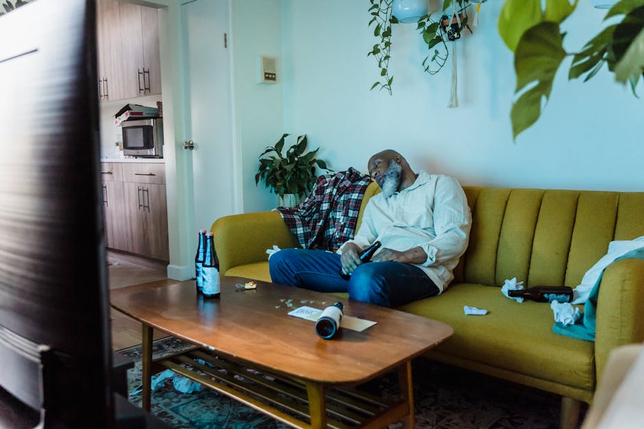 A man asleep on a yellow couch in a cozy living room with beer bottles and a TV remote.