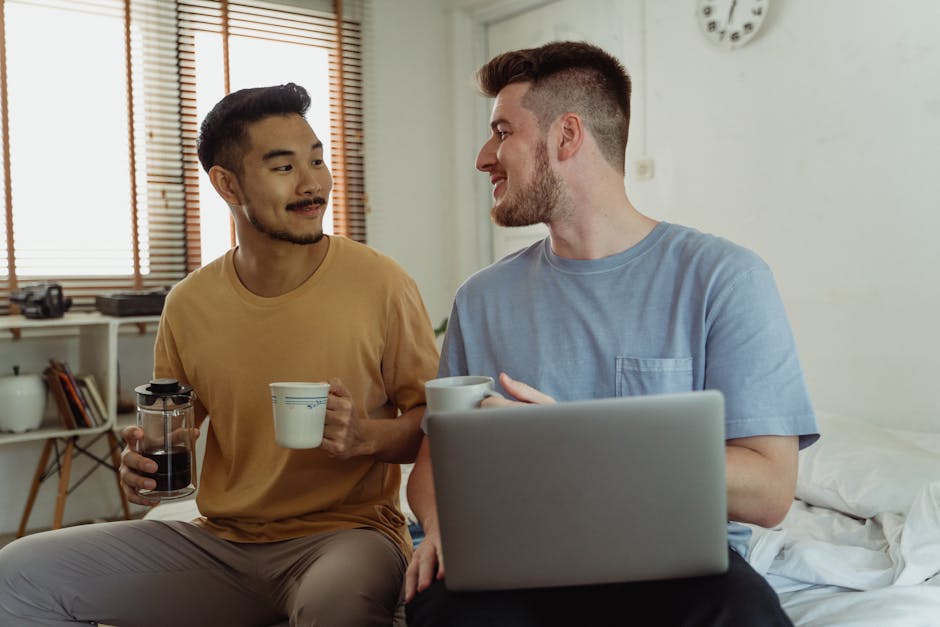 Two men sharing coffee and using a laptop in a cozy bedroom setting.