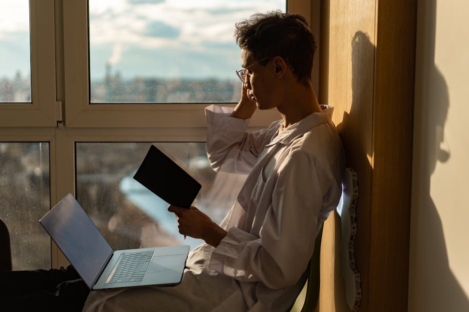 A young man reads beside a window with a laptop, enjoying the warm afternoon light.